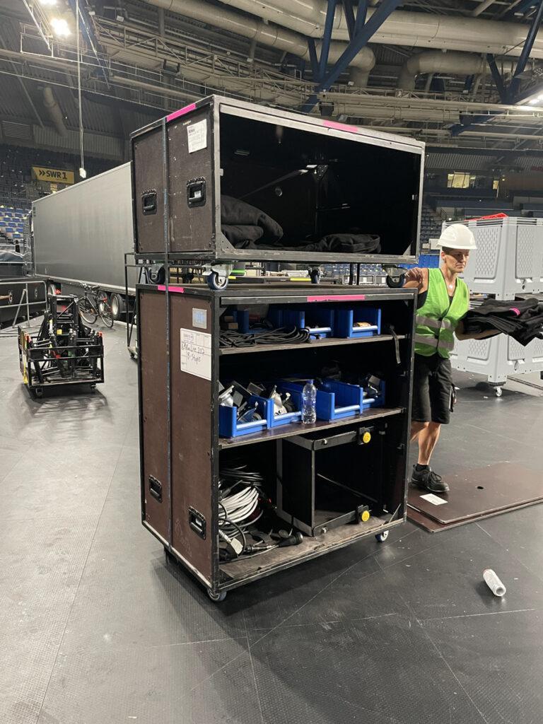 Zwei Cargo Carts in einer Eventhalle mit einer Frau daneben. Zwei Cargo Carts sind aufeinander gestapelt, offen an den Seiten und mit blauen Kisten sowie Kabeln gefüllt.